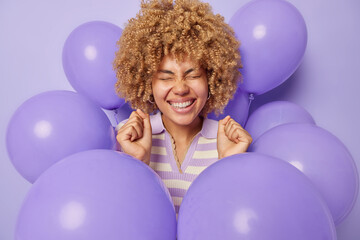 Fototapeta na wymiar Positive curly haired woman grins at camera smiles broadly clenches fists celebrates special occasion happy to receieve congratulations surrounded by inflated balloons isolated over purple background