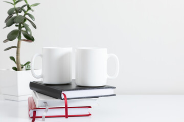 Two mugs mockup with notepad, accessories on white table and green plant in pot. Front view.