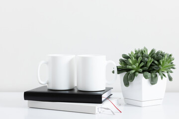 Two mugs mockup with notepad, accessories on white table and green plant in pot. Front view.