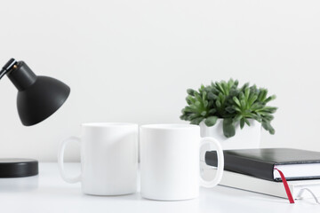Two mugs mockup with notepad, accessories on white table and green plant in pot. Front view.