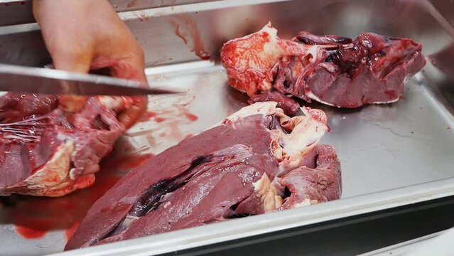 the butcher's bare hands carve a large bull's heart using a sharp large knife. A cut of a raw beef heart into pieces by a butcher in a deli. Offal of animals for human consumption. Meat industry