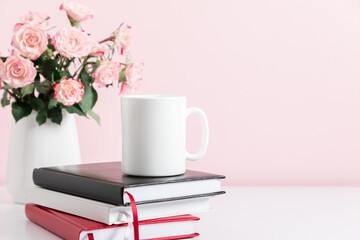 White cup mockup, stack of notebooks and bouquet pink roses in vase on pink table background. Front view. Place for text, copy space, mockup