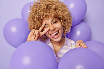 Naklejka premium Positive curly haired woman makes peace gesture over eye sticks out tongue has upbeat mood surrounded by inflated balloons isolated over purple background. Festivity and celebration concept.