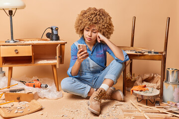 Fototapeta na wymiar Upset tired curly woman woodworker reads bad news via smartphone sits on floor takes break after fixes furniture surrounded by instruments and sawdust does carpentry work doesnt know what to do