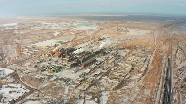 Flying a drone from a bird's eye view directly to the factory in a desert covered with snow and smoking chimneys