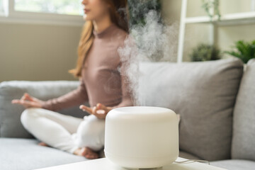 Fototapeta na wymiar Air humidifier, calm blurred woman, girl sitting on couch lotus pose put hands practice meditation do yoga exercise at home. Aromatherapy steam scent from essential oil diffuser in living room at home