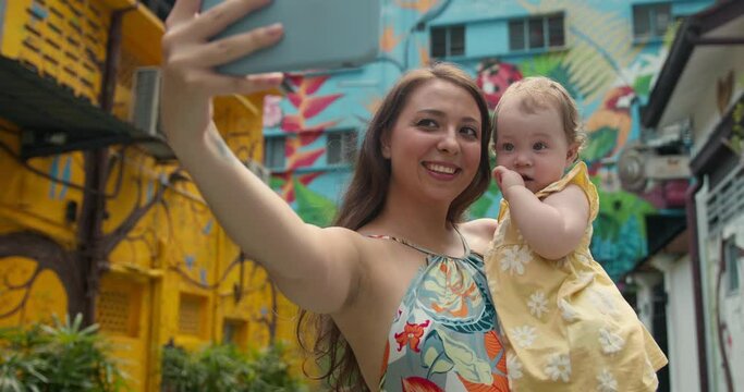Carefree smiling young mother and her adorable baby girl making selfie or video call to father or relatives at colorful background. Beautiful woman with daughter taking picture during holiday vacation