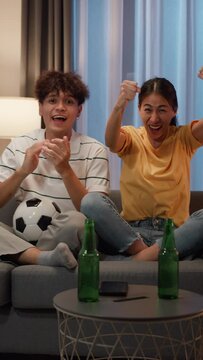 Asia couple sit on sofa excited cheering watching football sport game on television in living room at home. Young couple sport fans happy dancing in front of camera celebrate winner. Vertical Screen.