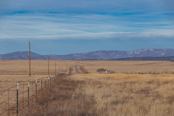 Barb wire fence and winding country road surrounded by yellow grass pastures with clear sky and mountain range in rural New Mexico