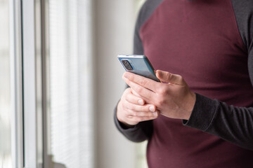 Close up of a man in sweater standing in front of windows at home and holding smartphone. Man hands holding smartphone