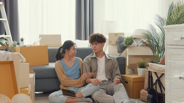 Young happy Asian couple sit on sofa couch use design thinking pointing wall get idea decoration room in living room at home. Husband wife move new house, Apartment Rent And Ownership Concept.