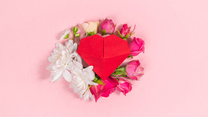 Red three-dimensional origami heart is decorated with natural flower buds. Pale pink background.