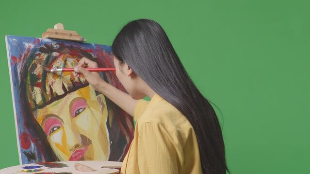 Side View Of Asian Female Artist Is Concentrated Painting A Girl On Canvas By Oil Paints And Brush In The Green Screen Studio
