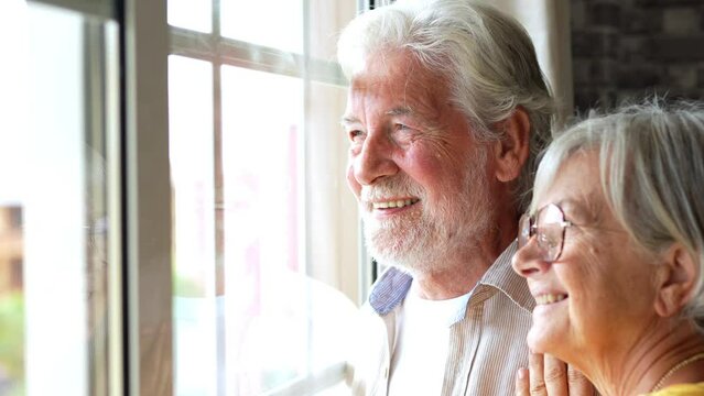 Happy Bonding Loving Middle Aged Senior Retired Couple Standing Near Window, Looking In Distance, Recollecting Good Memories Or Planning Common Future, Enjoying Peaceful Moment Together At Home.
