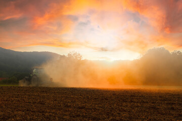 Tractor under cloudy dramatic sky from in red light of sunset sowing in plowed field.