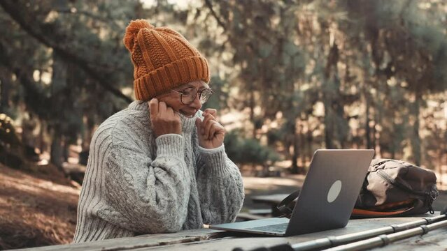 Head shot portrait close up of cute one of old middle age person using computer pc outdoors sitting at a wooden table in the forest of mountain in nature with trees around her.