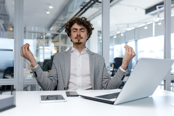 Hispanic businessman in bright white office relaxing at workplace, man meditating in lotus position at table with laptop, financier relaxing and breathing fresh air, boss in suit with eyes closed.