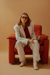 Fashionable confident woman wearing elegant white suit, trendy yellow glasses, leather ankle boots, sitting in armchair, posing on beige background. Full body studio fashion portrait
