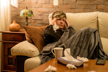 mujer mediana edad enferma con dolores  en el sofa de casa tapada con una manta, tomando medicina...
