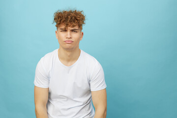 sad, gloomy young man with curly hair stands on a light blue background and looks into the camera, emotionally maning his face with disappointment. Photo with space for inserting an advertising layout © Tatiana