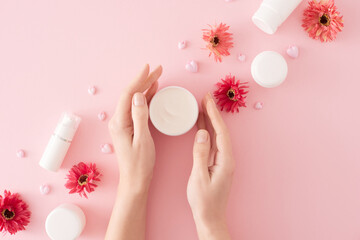 Women skin care concept. Flat lay photo of cream jar in hands, heart shaped candles, cosmetic bottles and flowers on pastel pink background. Mother's day cosmetic idea.