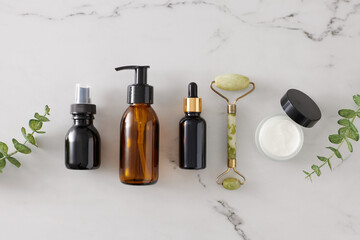 Skincare products concept. Top view photo of amber cosmetic bottles, cream jar, dropper bottle, face massage roller and eucalyptus leaves on marble background. Flat lay cosmetic mockup.