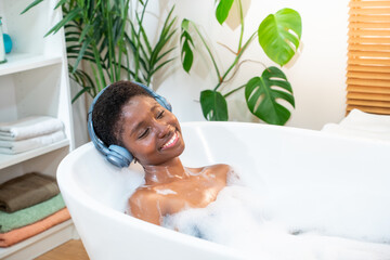 Smiling African attractive woman wearing headphones relaxing with music, podcast or audio book in foam bath in bathroom. Beauty, skin care, home spa and wellbeing concept. High quality photo