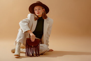 Fashionable confident woman wearing elegant white suit, hat, leather ankle boots, holding brown suede fringed bag, sitting, posing on beige background. Copy, empty space for text
