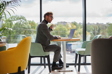 Full-length portrait of a businessman drinking coffee while telecommuting on laptop in modern office