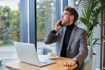 Young businessman talking on mobile phone, sitting at desk with laptop by a cup of aroma coffee