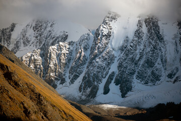 Peaks of stone mountains with tongues of glaciers and snow are covered with white clouds and fog in...