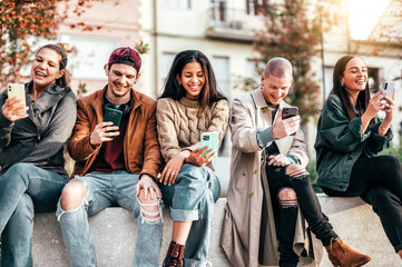 Teenagers texting mobile phone messages sitting on banche - Group of young multiracial friends...