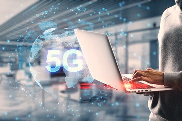 Close up of hands using laptop with globe and 5g hologram on blurry office interior background. Telecommunication and internet speed concept. Double exposure.