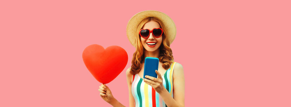Portrait of happy smiling young woman with smartphone and red heart shaped balloon wearing summer straw hat on pink background, blank copy space for advertising text