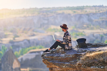 Man traveler blogger works remotely at computer sitting on a cliff at calm destination. Work...