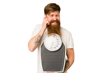 Adult redhead man with long beard holding a scale isolated cut out covering ears with hands.