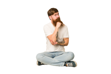 Young adult redhead with a long beard sitting on the floor isolated thinking and looking up, being reflective, contemplating, having a fantasy.
