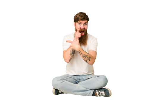Young adult redhead with a long beard sitting on the floor isolated saying a gossip, pointing to side reporting something. - Powered by Adobe
