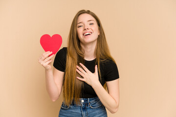 Young redhead woman holding a paper red heart cut out isolated laughs out loudly keeping hand on chest.