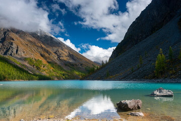 Stones lies in the water near the shore of the turquoise mountain lake Shavlinsky between stone rocks with fog and clouds and forest in Altai.
