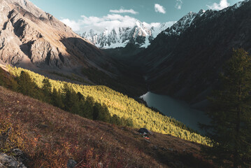 Panorama Mountain lake Shavlinskoe in the shade without sun between the rocks behind the forest against the backdrop of snowy peaks with glaciers in Altai.