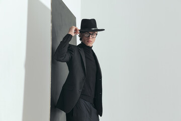 cool young guy with glasses adjusting hat while posing in front of black wall