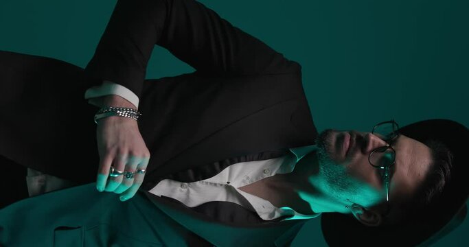 vertical video with colorful light of best man with undone bowtie and hat adjusting tuxedo and looking to side, holding hands in pockets