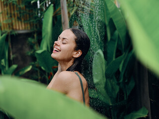 A woman takes a shower and washes her head and hair outdoors, closed eyes and a smile on the background of tropical plants, palm trees, green banana leaves, summer rain