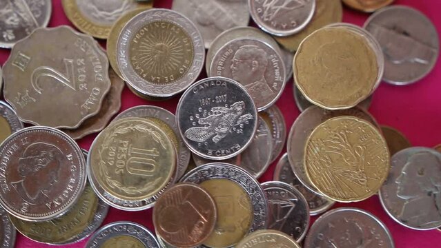 Coins from different currencies around the world are droped and staked on a  colorful pink surface 