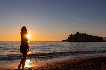 Fototapeta premium Silhouette of barefoot woman on sand beach with scenic romantic view on idyllic island Sveti Stefan at sunset, Budva Riviera, Adriatic Mediterranean Sea, Montenegro, Europe. Summer vacation at seaside