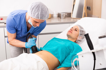 Confident male cosmetologist performing body ultrasound cavitation procedure for young man at aesthetic clinic