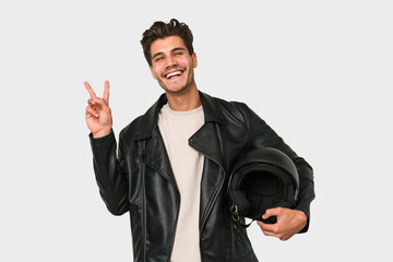 Young caucasian man holding a motrbike black helmet isolated joyful and carefree showing a peace...