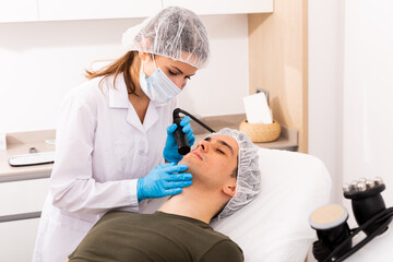 Young man receiving face rejuvenation treatment on modern equipment at cosmetology clinic, male skincare concept