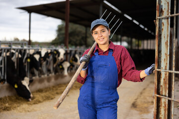 Young female farmer in overalls posing while feeding cows at dairy farm